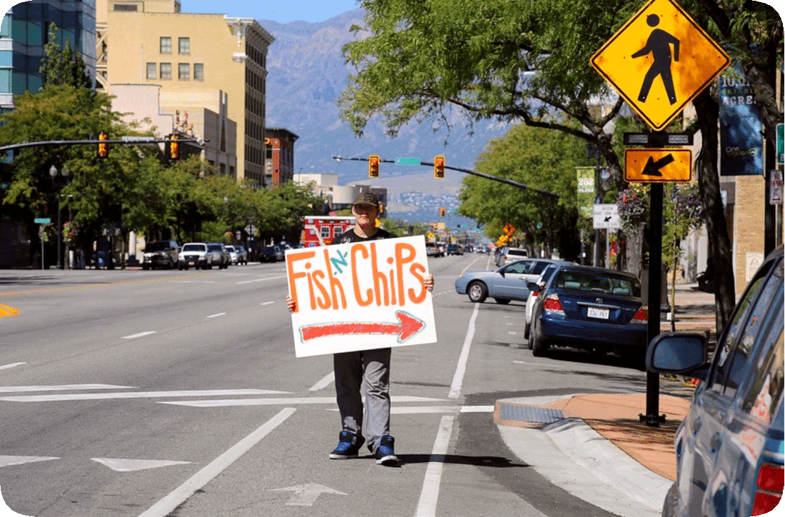 Picture of a man holding a sign reading ‘Fish and Chips’ with an arrow on it pointing to a restaurant location.