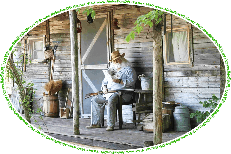 Picture of a man with a beard and shoulder-length white hair sitting on a chair on the porch of a small rustic wooden house in a woods, reading a book, with a double-barreled shotgun resting across his lap, and the words, ‘Visit www.MakeFunOfLife.net.’