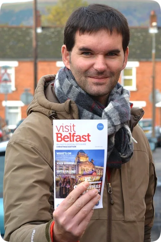 Picture of a happy smiling Irishman holding up a travel brochure with the printed words, 'Visit Belfast.'